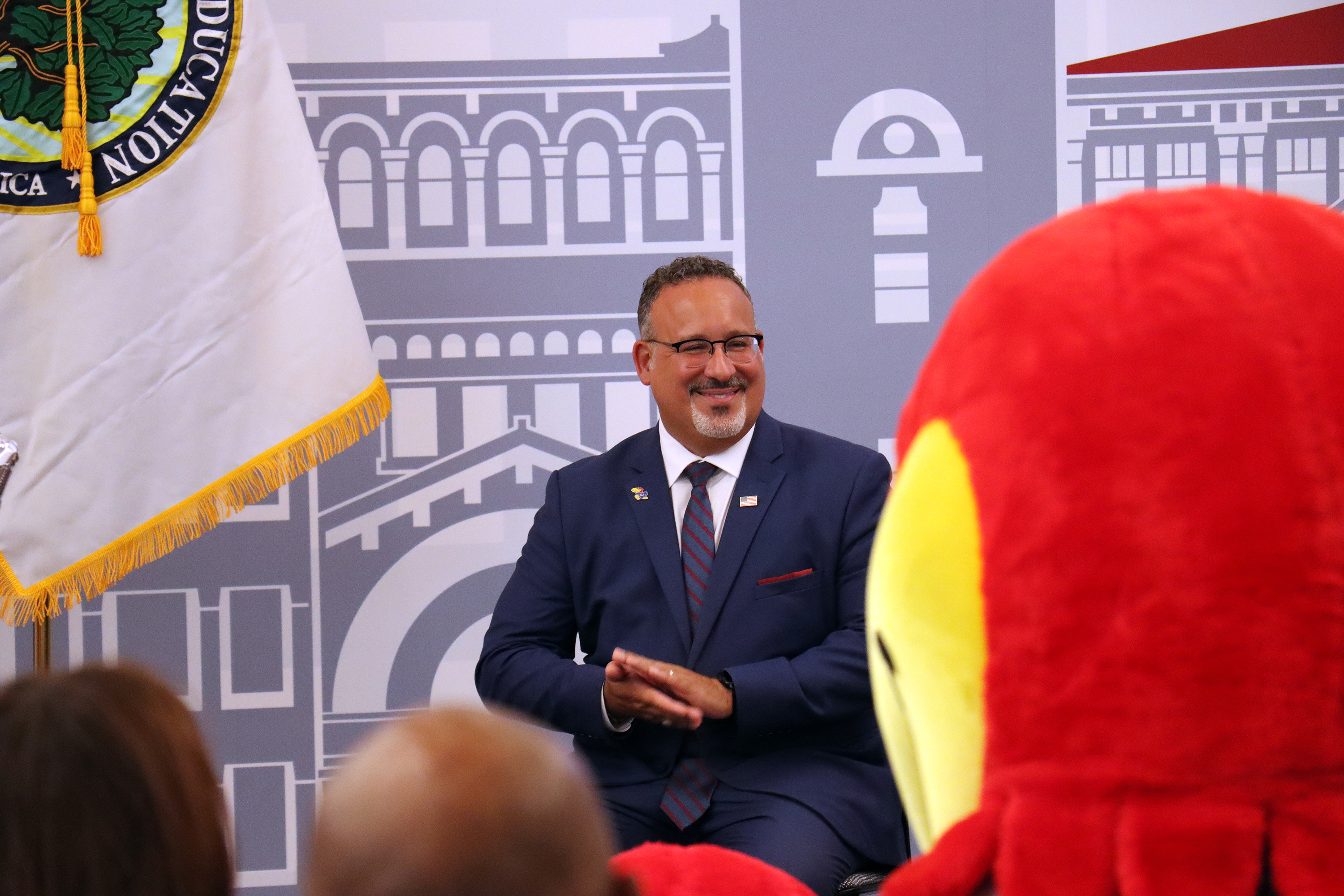  U.S. Secretary of Education Miguel Cardona sits, foregrounded by the heads of attendees and KU mascot Big Jay. 